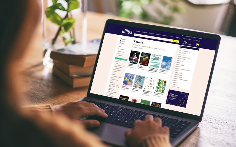 En person använder en bärbar dator med Ellibs nätbokhandel öppen på skärmen. I bakgrunden syns böcker och en grön växt på bordet.