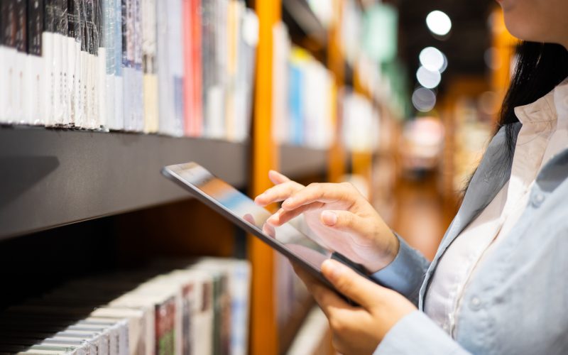 A person using a tablet while standing by a bookshelf in a library.