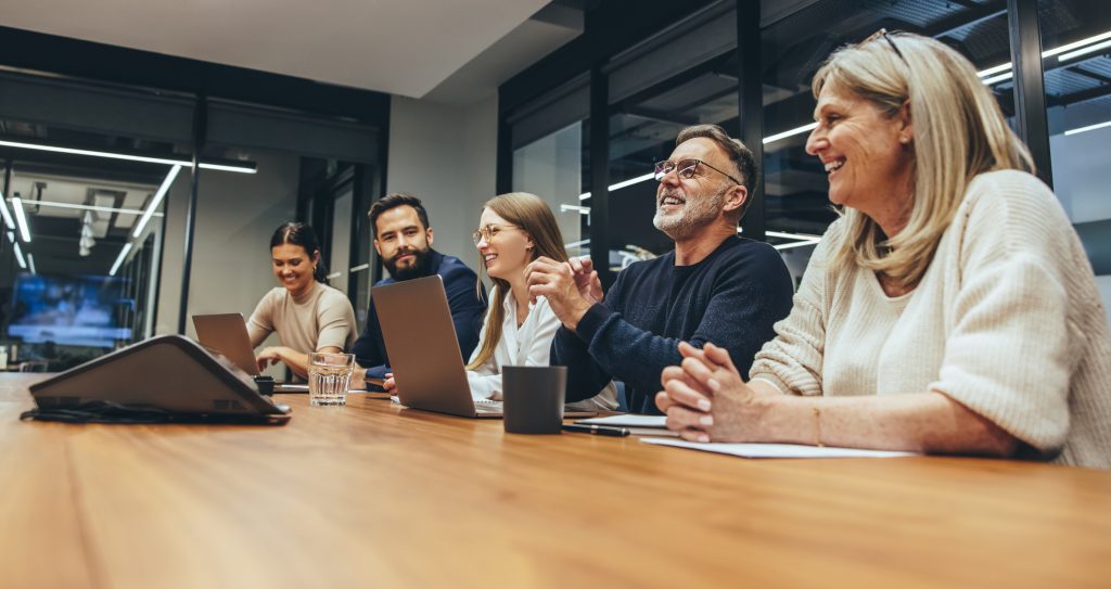 A group of people of different ages sitting around a long conference table, smiling and talking during a meeting. Several have laptops in front of them.