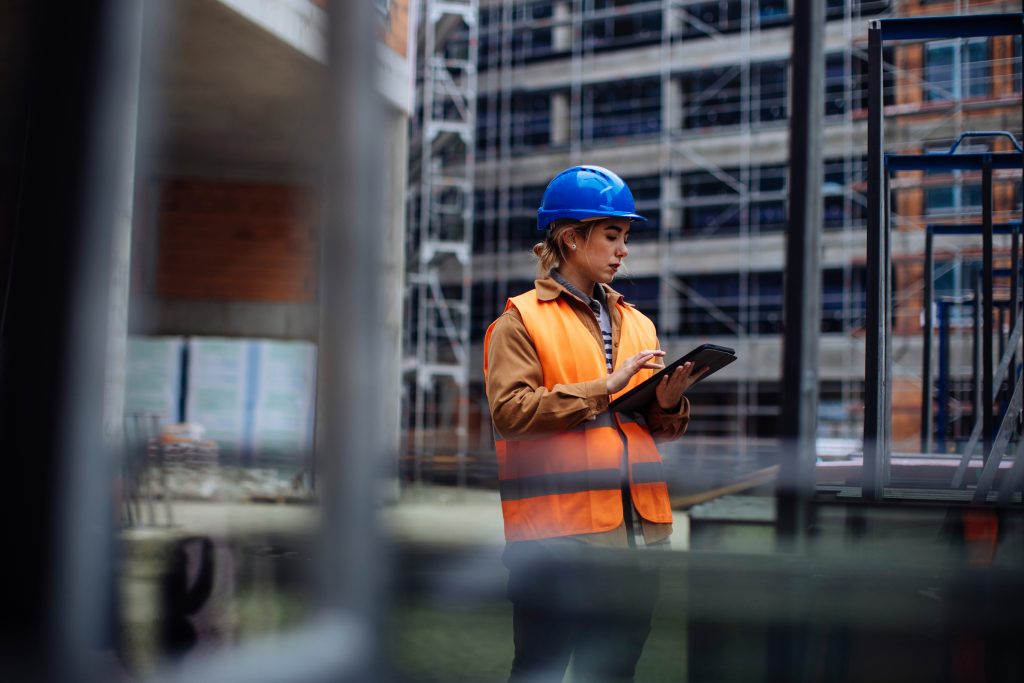A person on a construction site wearing a safety vest and a blue hard hat is using a tablet. The background shows the building structure and scaffolding.