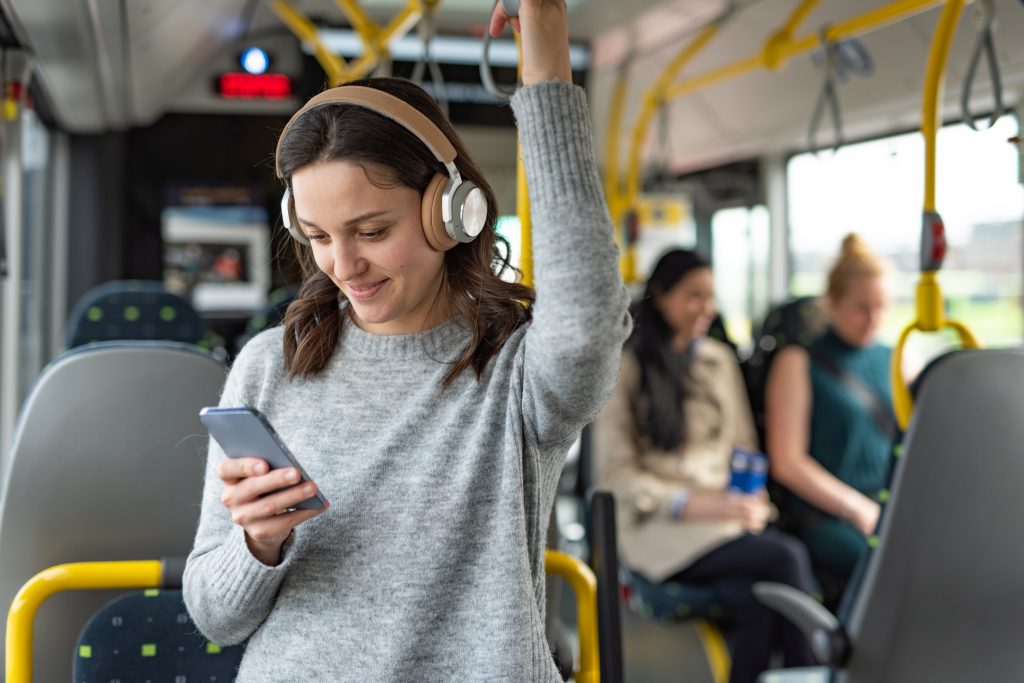 A young woman standing on a bus wearing headphones, holding onto a handrail and smiling at her phone. Other passengers are visible in the background.
