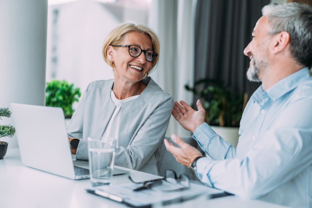 Två kollegor sitter vid ett bord med en bärbar dator och pratar glatt med varandra.