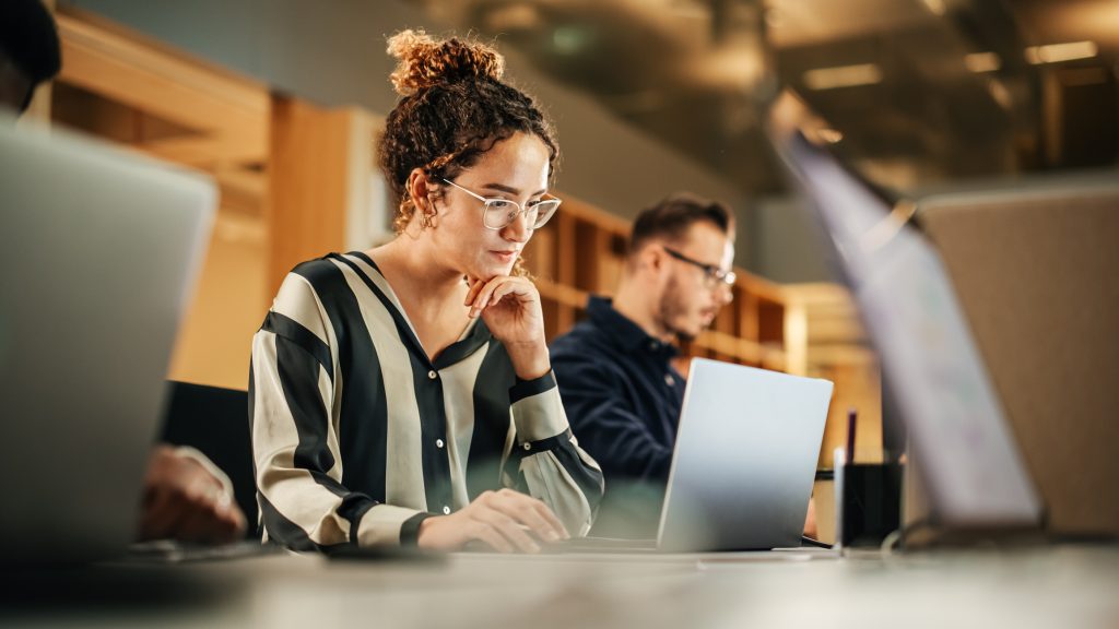 A woman working intently on a laptop in an office environment, with colleagues in the background.