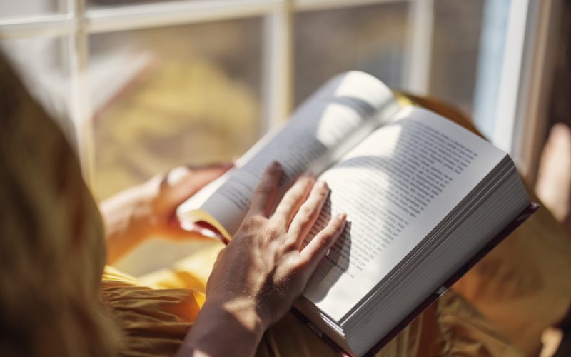 Henkilö lukemassa kirjaa kirkkaan ikkunan ääressä, auringonvalo luo pehmeitä varjoja avoimille sivuille.