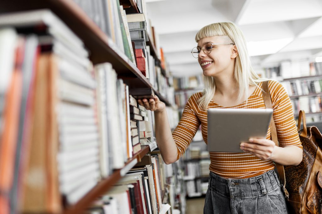 Young female student smiling while searching for a book on a library shelf, holding a tablet in her hand.