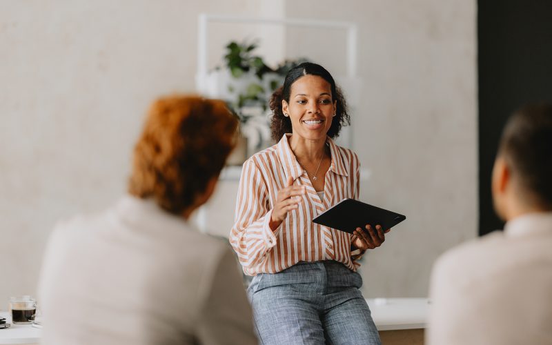 En leende person håller en presentation för en liten grupp och håller en surfplatta i handen. Två deltagare syns i förgrunden med ryggen mot kameran.