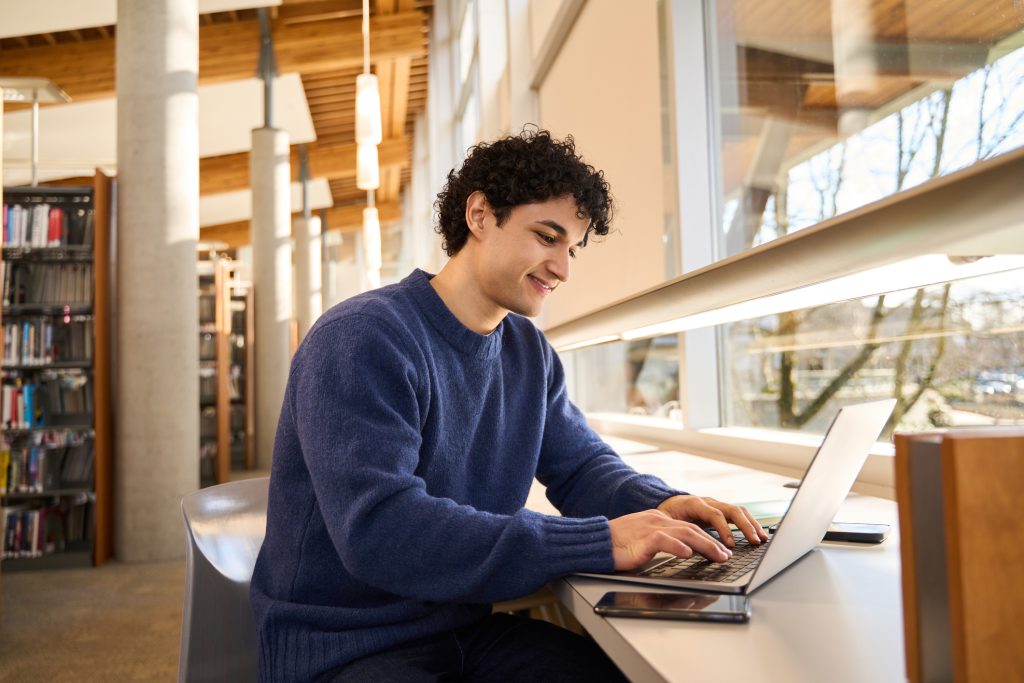 A young man working on a laptop by a window in a library, with natural light streaming in.