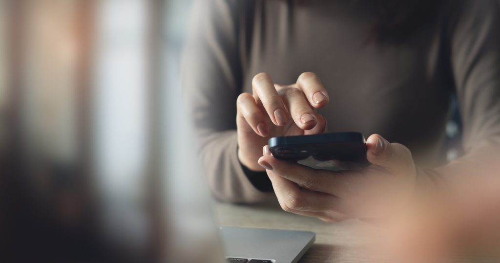 A person using a smartphone at a table with a laptop visible in the background.