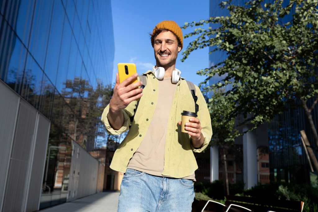 A man walking outdoors in an urban setting, smiling while looking at his yellow phone and holding a coffee cup.