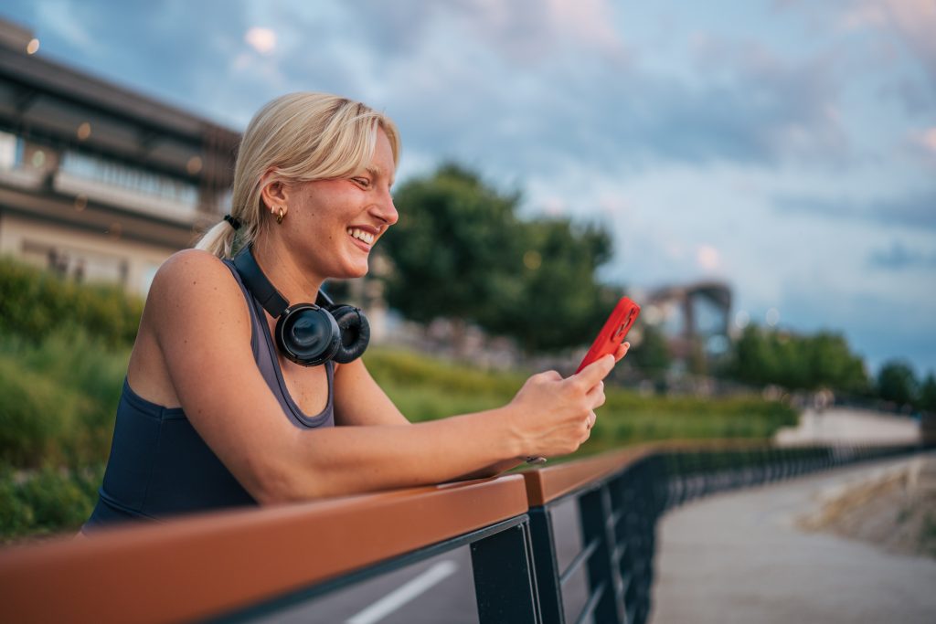 En kvinna lutar mot ett räcke utomhus, ler och tittar på sin röda telefon, med hörlurarna runt halsen.