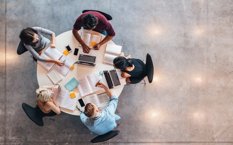 Top-down view of a group of people working together at a round table with books, notebooks, and laptops.