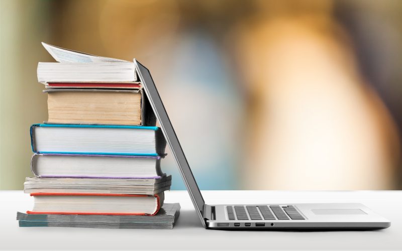 A stack of books on a desk next to an open laptop, with a softly blurred, warm-toned background.