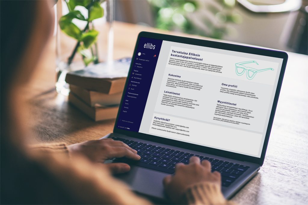 Henkilö käyttää kannettavaa tietokonetta, jonka näytöllä on auki Ellibsin kustantajapalvelu. Taustalla näkyy kirjoja ja vihreä kasvi pöydällä.