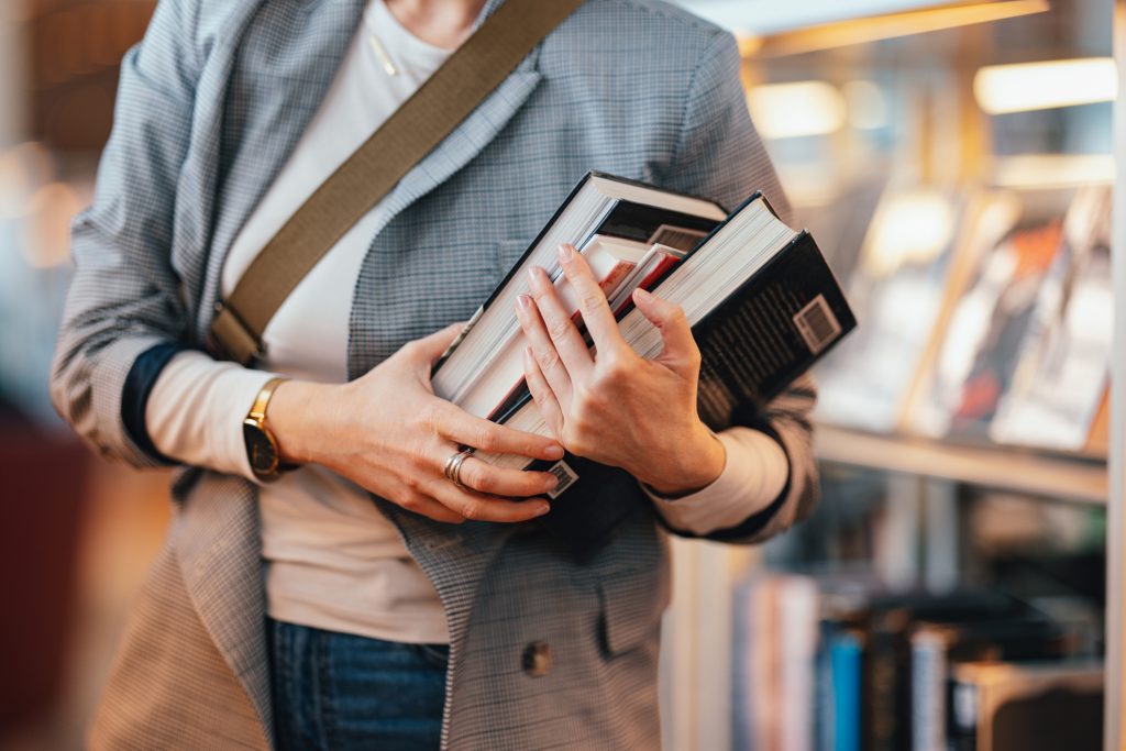 A person holding a stack of books in their arms while standing in a bookstore, dressed in a blazer and casual clothing.